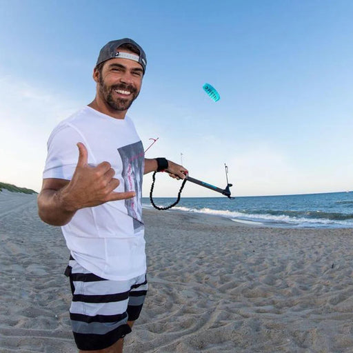 Man holding wing foil on beach, wearing white tee and striped shorts, kite flying overhead