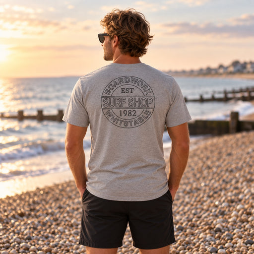 Man wearing the Boardworx Surf Shop grey T-shirt on a Whitstable-style shingle beach, back logo view