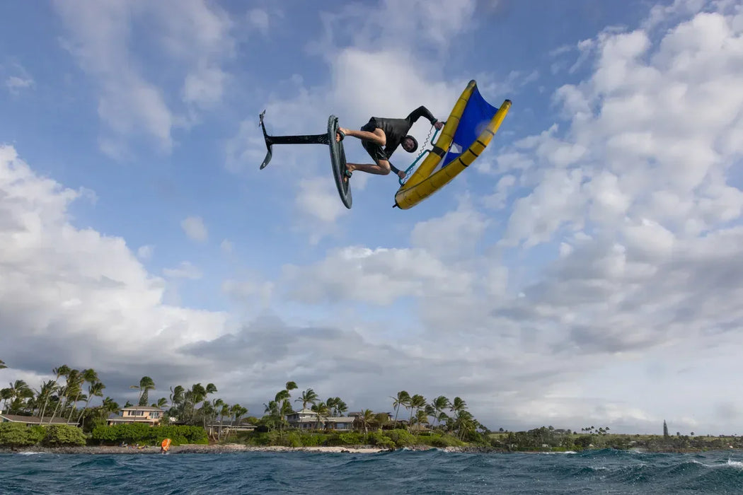 Man performing aerial trick on carbon foil mast wing foil with yellow and blue wing, coastal backdrop
