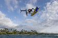 Man performing aerial trick on carbon foil mast wing foil with yellow and blue wing, coastal backdrop