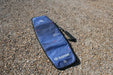 Duotone blue twin tip board bag with shoulder strap on a pebbled beach, 143cm size visible.
