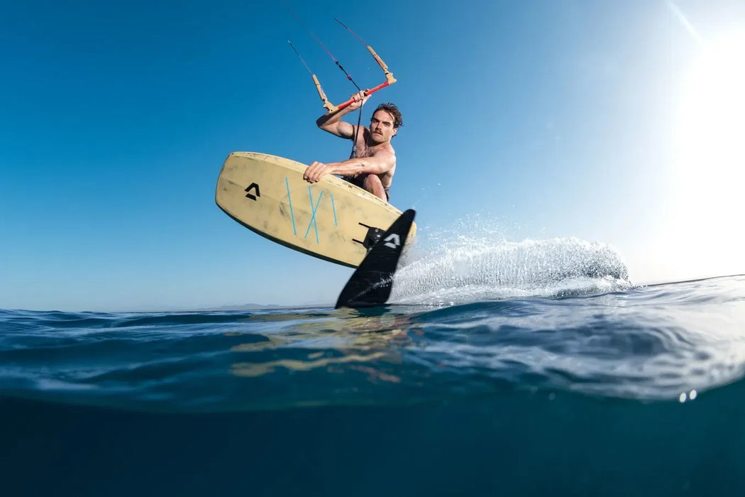 Man kitesurfing on Duotone foil board, tan and black, over blue water under clear sky