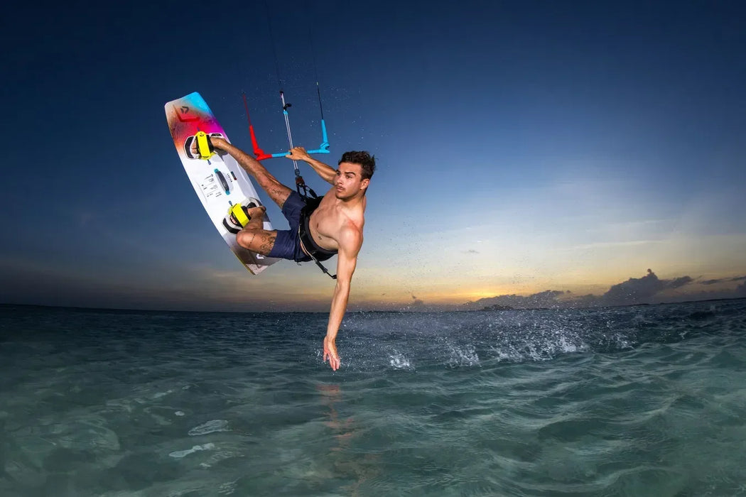 Duotone twin tip kiteboard with yellow foot straps in action, kitesurfer jumping at sunset over clear water.