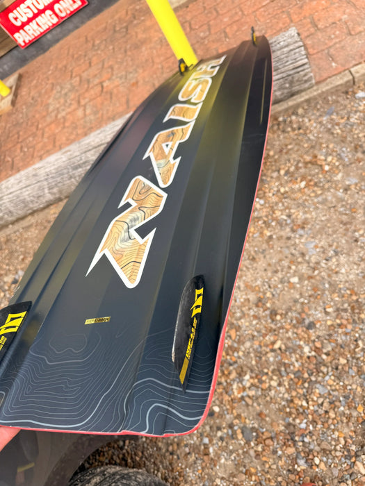 Naish Monarch kitesurfing board, black with wood grain logo and yellow fins, on gravel