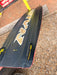 Naish Monarch kitesurfing board, black with wood grain logo and yellow fins, on gravel