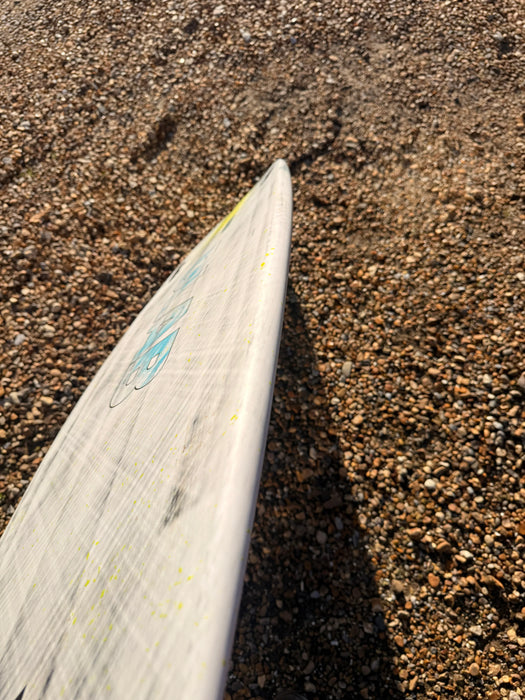 Duotone Whip D/LAB white surfboard on pebbled beach, close-up of board edge and logo