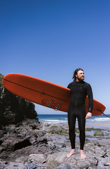 Man in black chest zip wetsuit holding orange surfboard on rocky beach