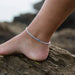 White beaded anklet with blue center bead on ankle, worn outdoors on rocky beach